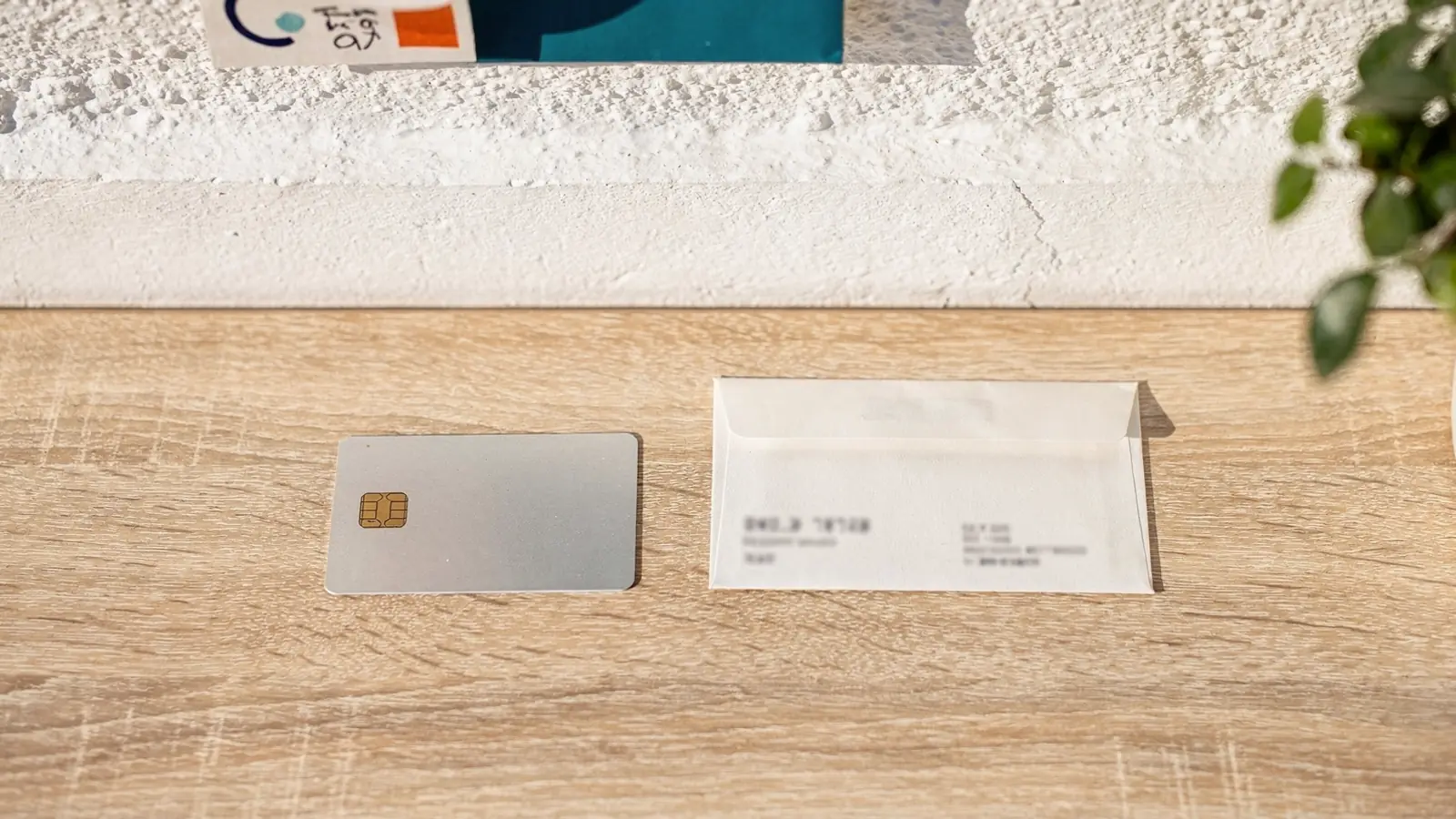 Una tarjeta bancaria plateada con chip dorado y un sobre de papel blanco sobre una mesa de madera clara, con luz natural.